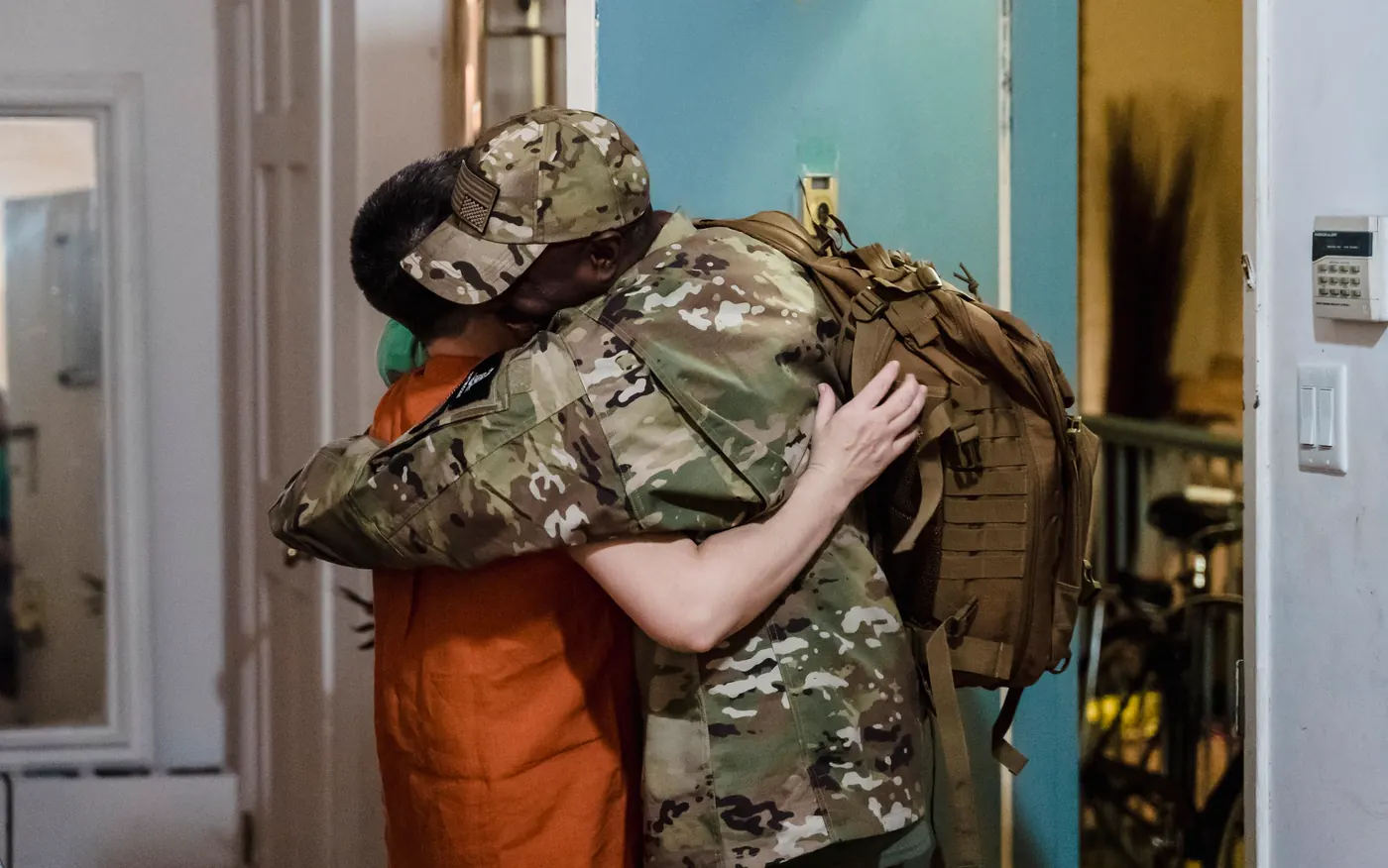 Generic stock photo: a soldier in camouflage with a backpack hugging a family member at a front door